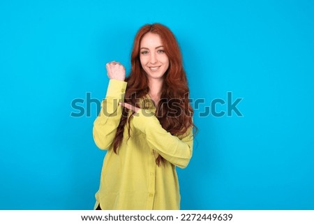 young beautiful red haired woman wearing green shirt over blue studio background In hurry pointing to wrist watch, impatience, looking at the camera with relaxed expression