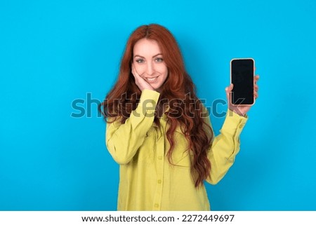 young beautiful red haired woman wearing green shirt over blue studio background hold hand modern technology use touch face palm astonished impressed scream wow omg unbelievable unexpected