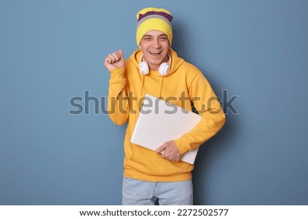 Photo of overjoyed smiling cheerful man wearing beanie hat and casual yellow hoodie, holding closed laptop, clenched fist, finishing project, celebrating, posing against blue wall