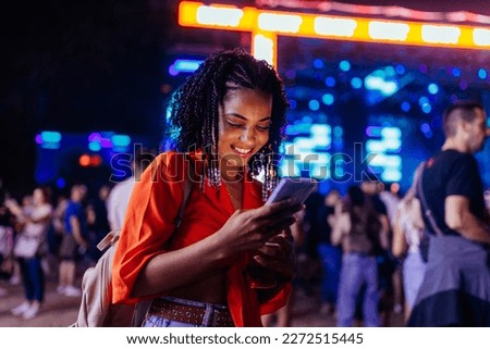 A young African American girl is with a smartphone at a music festival, texting with her friends. Royalty-Free Stock Photo #2272515445