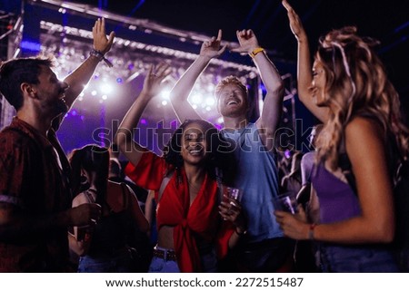A young crowd of multiethnic friends are dancing at a concert having beers and holding their hands up in their casual clothing. Royalty-Free Stock Photo #2272515487