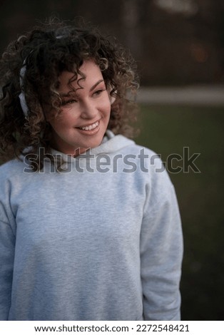 Young curly haired woman in grey sporty suit in headphones has fun outdoors in the evening