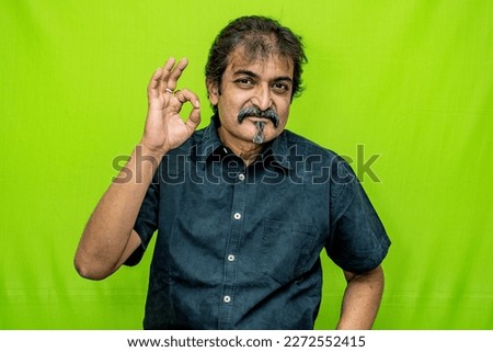 The well-dressed Indian man, wearing a black shirt, is showing appreciation with an appropriate sign using his right hand's forefinger and thumb while standing against a green screen background
