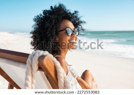 Happy young black woman relaxing on deck chair at beach wearing spectacles. Smiling african american girl with sunglasses enjoy vacation. Carefree happy young woman sunbathing at sea with copy space. Royalty-Free Stock Photo #2272553083
