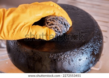 A man's hand in a protective glove cleans a pan with a burnt bottom using a metal kitchen sponge. Royalty-Free Stock Photo #2272553451