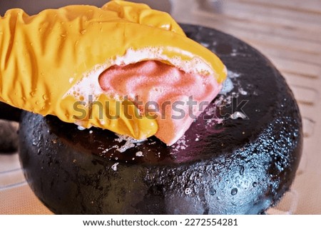 A man's hand in a protective glove cleans a pan with a burnt bottom using a foam kitchen sponge. Royalty-Free Stock Photo #2272554281