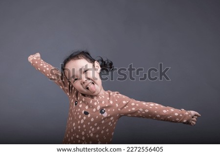 portrait on a gray background of a little cheerful Asian girl with her tongue hanging out and her arms to the sides in a brown longsleeve