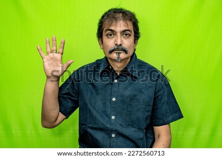 The well-dressed Indian man wearing a black shirt is indicating the number 'five' with his right hand's two fingers while standing against a green screen background