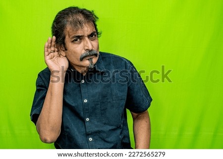 The well-dressed Indian man, dressed in a black shirt, stands against a green screen background and is seen intently cupping his right ear as if trying to hear a quiet or secret sound