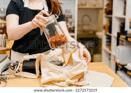 Close up of sales assistant standing behind the counter in sustainable plastic free wholefood store holding container of goji berries. Royalty-Free Stock Photo #2272573219
