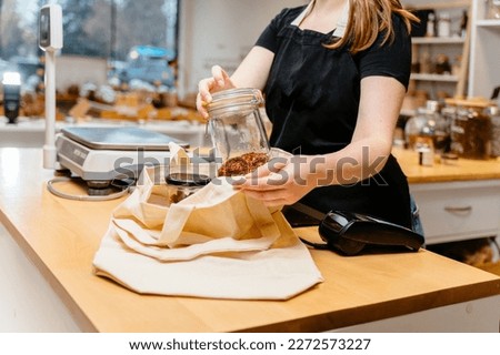 Close up of sales assistant in sustainable plastic free wholefood store holding container of goji berries, weighing products goods in glass containers in local zero waste grocery store. Royalty-Free Stock Photo #2272573227