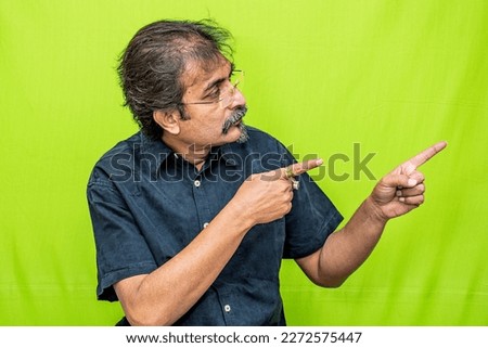 The well-dressed Indian man wearing a black shirt is pointing in the right direction with both of his hand's fingers while standing against a green screen background