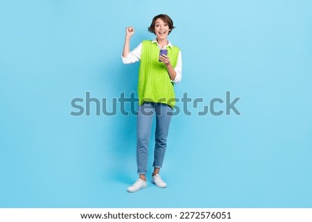 Full size photo of impressed girl with brown hairdo wear green vest raising fist scream yes hold phone isolated on blue color background