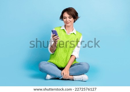 Full size photo of cheerful girl with brown hairdo wear green vest jeans look at phone read email isolated on blue color background