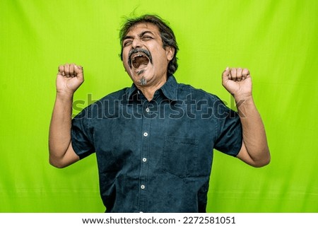 An Indian man, dressed in a sharp black shirt, is standing in front of a green screen. He yawns and relaxes, lifting both fists up to his shoulders