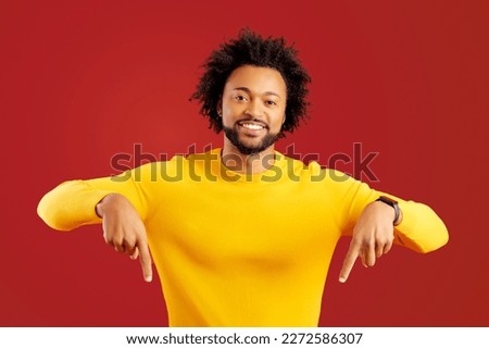 Here and now. Bearded handsome man pointing finger down, looking at camera with happy facial expression. Indoor studio shot isolated on red background Royalty-Free Stock Photo #2272586307