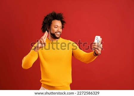 Portrait of young african american handsome man taking selfie, looking at camera and gesturing hand, curly guy recording himself. Indoor studio shot isolated on red background