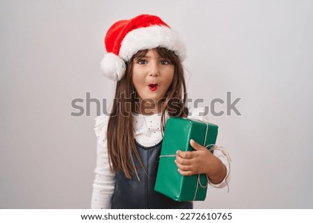 Little hispanic girl wearing christmas hat and holding gifts scared and amazed with open mouth for surprise, disbelief face 