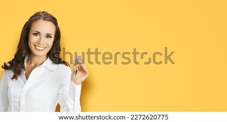 Happy smiling young businesswoman or real estate agent showing keys from new house. Portrait of brunette business woman, isolated on orange yellow background.