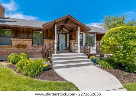 Facade of home with paved walkway leading to the portico and white front door. Front yard with lush foliage and flowers, stone wall, sidelight, and outdoor stairs can also be seen at the exterior. Royalty-Free Stock Photo #2272652835