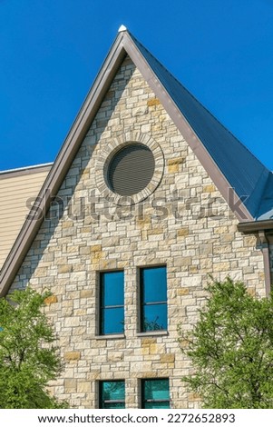 Vertical shot of a french country home with round attic window and reflective windows- Austin, Texas. There are trees outside near the reflective tall windows of the house with stone veneer sidings. Royalty-Free Stock Photo #2272652893