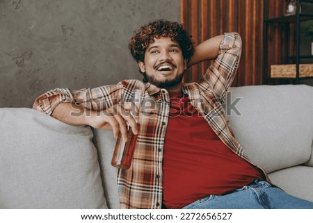 Young smiling fun Indian man wears casual clothes hold bottle of beer look aside sits on grey sofa couch stay at home hotel flat rest relax spend free spare time in living room indoor. Lounge concept