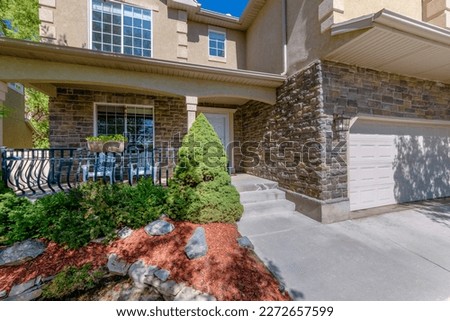 Two-storey house with stone veneer siding and attached garage. House entrance with plants on the left near the porch with planter on railings and white chairs near the windows beside the white door. Royalty-Free Stock Photo #2272657599