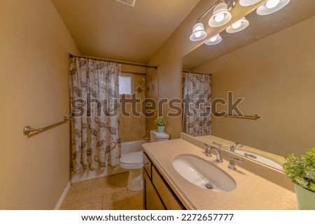 Bathroom interior with yellowish light brown walls and flooring tiles. There is a bar handle on the left near the printed shower curtain on the tub shower wint window and vanity sink on the right. Royalty-Free Stock Photo #2272657777