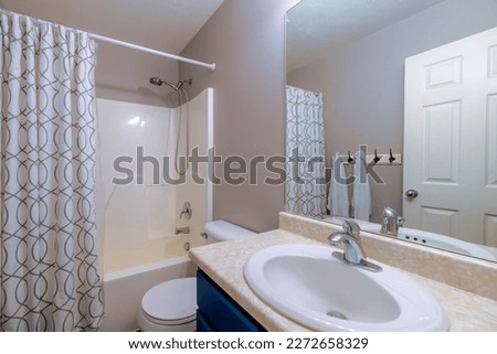 Small bathroom interior with light gray wall and acrylic tub shower. There is a sink on the right with mirror and reflection of white door and hanging towels across near the shower curtain of the tub. Royalty-Free Stock Photo #2272658329