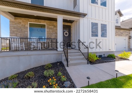 House entrance with pathway lights on the lawn at the front heading to the porch with railings. Home exterior with stone veneer and white board and batten siding near the black front door with wreath. Royalty-Free Stock Photo #2272658431