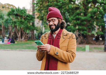 Happy bearded male hipster typing messages with a mobile phone social media app at city park outdoors. Portrait of young caucasian man using a smartphone to watch funny content. Technology concept