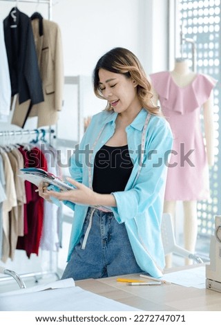 Millennial Asian young cheerful professional female dressmaker designer seamstress standing smiling working alone holding looking at drawing sketching paper collection in tailor workshop studio office
