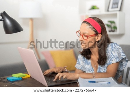 Young woman with red glasses and pink headband, using her pink laptop to write a college paper, sitting at the desk in her apartment.