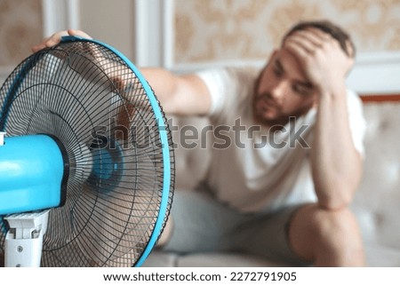 Young bearded man using electric fan at home, sitting on couch cooling off during hot weather, suffering from heat, high temperature Royalty-Free Stock Photo #2272791905