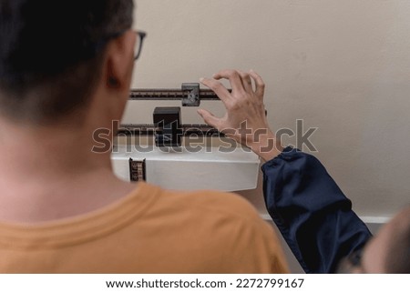 A male patient is being weighed with a physician beam scale at a local clinic. Adjusting the smaller counter weight until the beam rests horizontally. Royalty-Free Stock Photo #2272799167