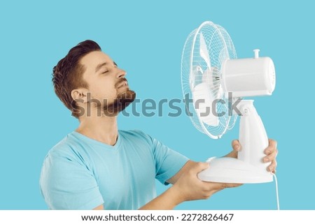 Man using an electric fan to survive the summer heat. Happy guy standing with his eyes closed on a blue background, holding a white fan in his hands, and enjoying cool, fresh air blowing in his face Royalty-Free Stock Photo #2272826467