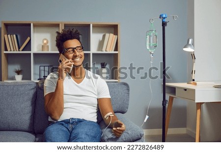 Happy, smiling young African American man talking with doctor on cell phone while sitting on couch at home and receiving medication infusion through intravenous drip Royalty-Free Stock Photo #2272826489