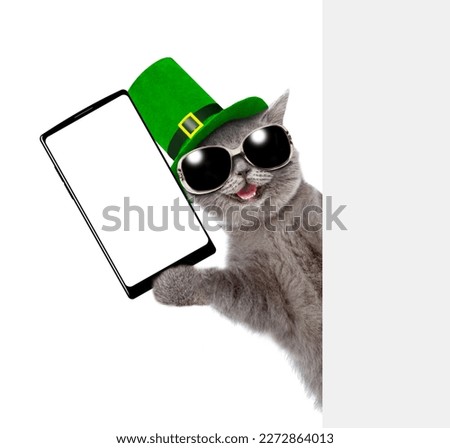 St. Patricks Day. Happy cat wearing sunglasses and green hat of the leprechaun holding big smartphone with white blank screen. isolated on white background