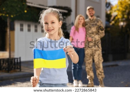 Child with Ukrainian flag, her father in military uniform and mother on city street, space for text. Family reunion