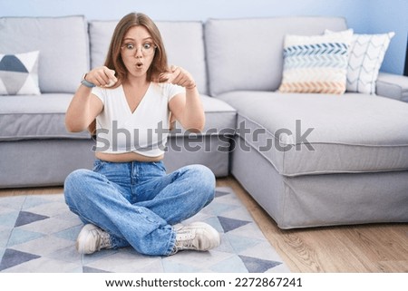 Young caucasian woman sitting on the floor at the living room pointing down with fingers showing advertisement, surprised face and open mouth 
