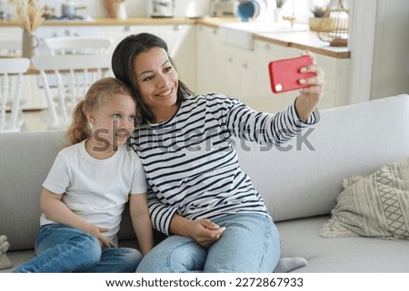 Happy mom and little daughter takes home selfie together, sitting on sofa, holding smartphone, looking at phone screen. Smiling mother and kid girl makes video call, chatting online, using mobile app.