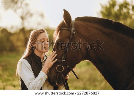 Young beautiful female jockey strokes and hugs the horse's head and prepares for the competition. Jumping training in the meadow in summer evening. Royalty-Free Stock Photo #2272876827