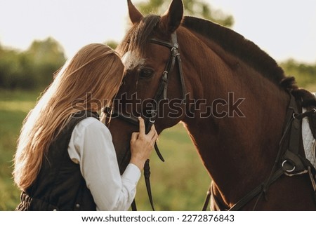 Young beautiful female jockey strokes and hugs the horse's head and prepares for the competition. Jumping training in the meadow in summer evening. Royalty-Free Stock Photo #2272876843