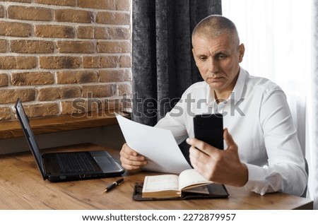 A businessman holds a phone and documents in his hands, reads information from the phone. Financial manager, company.