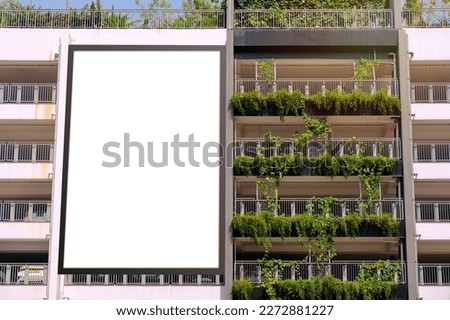 Blank white vertical advertising billboard banner mockup, outside multi-storey carpark with lush green plants. Large digital display screen poster, an out-of-home OOH media display space.