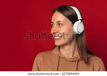 Pretty smiling woman is wearing headphones and looking aside at copy space over red background.