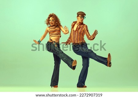 Dynamic portrait of happy man and woman in retro colored shirts and flared jeans dancing energetic dance over green background. Concept of fashion trends of 70s, 1980s years, music, hippie lifestyle Royalty-Free Stock Photo #2272907319