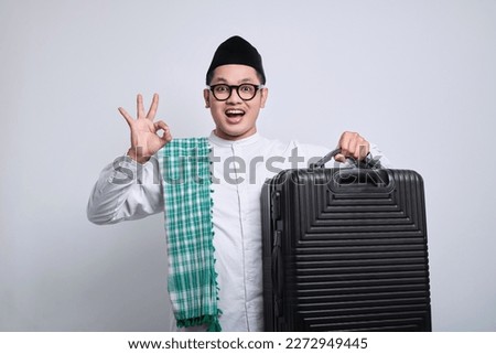 Excited young Asian Muslim man in white prayer clothes standing over isolated white background showing ok sign while holding a black suitcase.