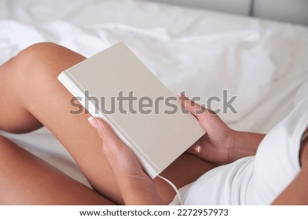 Young woman holding blank notebook or book mockup with white cover while sitting on the bed.