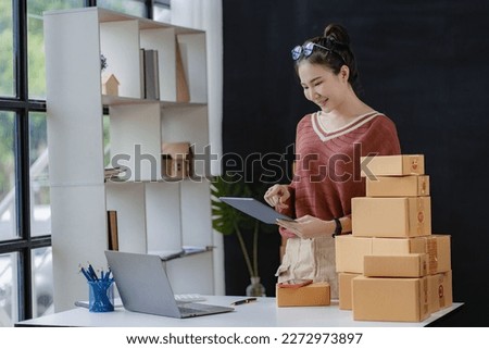 Asian female business owner using laptop to take and check online orders to prepare boxes. Packing products for delivery to customers, SME business concept, e-commerce, online shopping